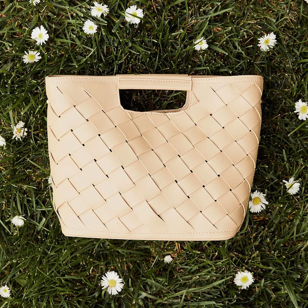 a beige woven handbag on the grass with daisies surrounding it