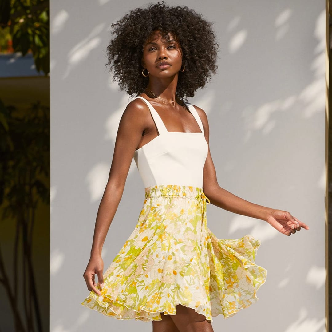 a woman walking outside wearing a white tank top and a green and yellow 70s floral print mini skirt