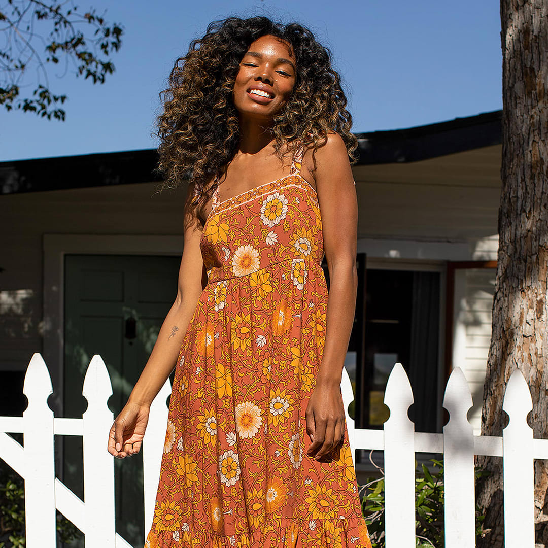 a model in front of a white fence wearing a rust brown floral print tie strap maxi dress