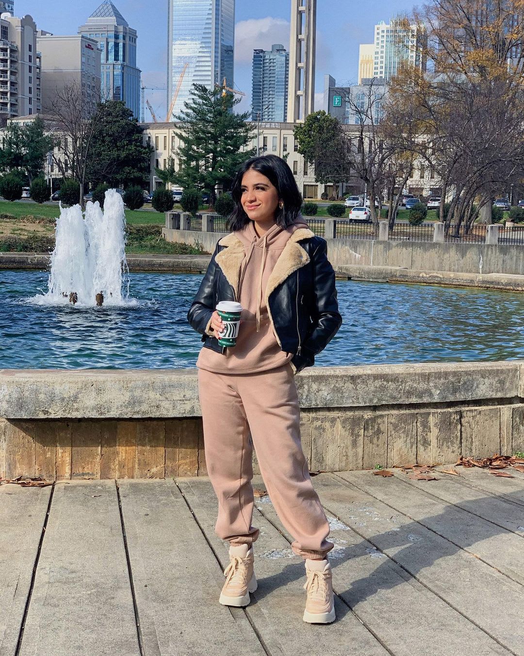a woman in a sweatsuit wearing a shearling jacket standing in front of a fountain looking into the distance of her community