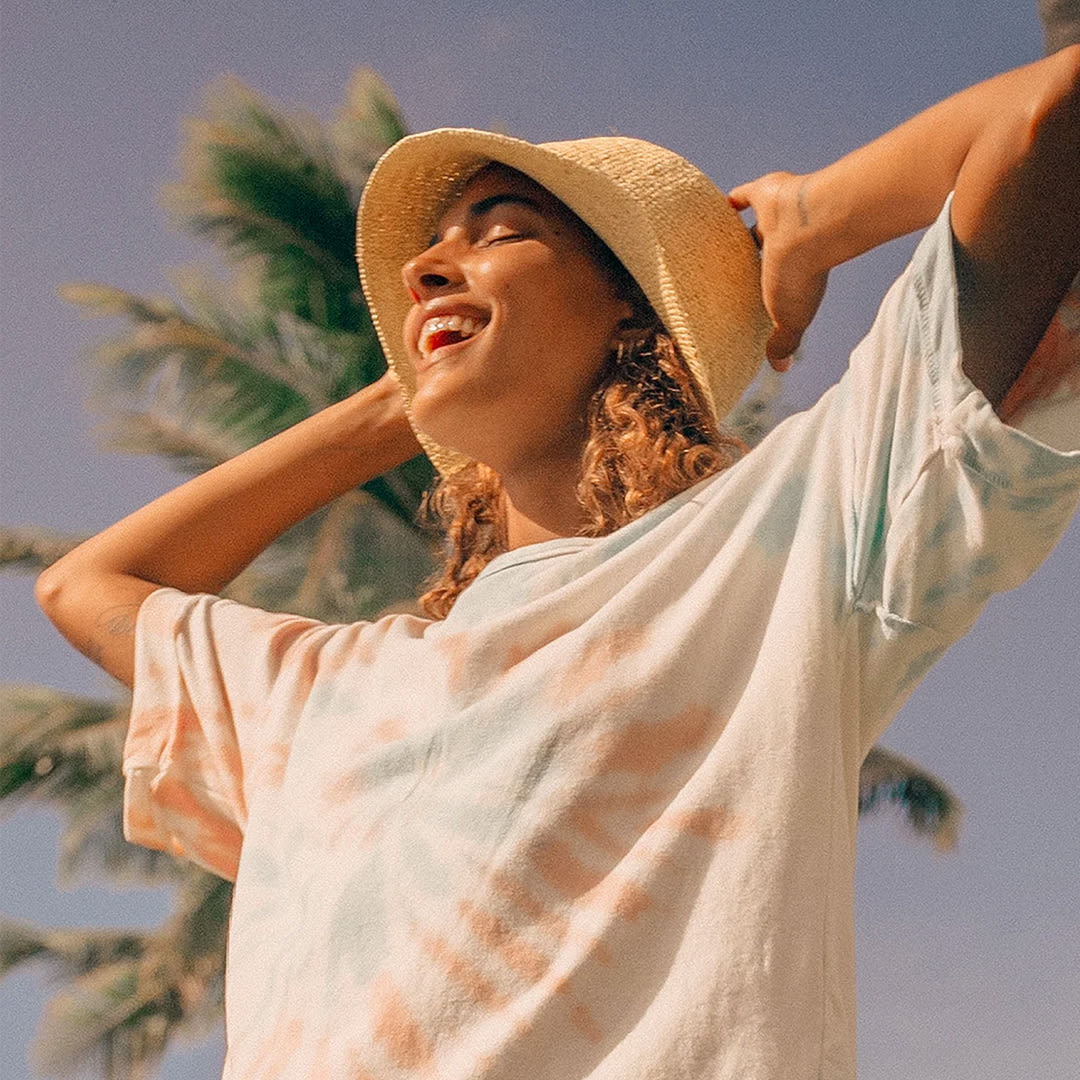 a woman wearing a tie dye tee and straw bucket hat, smiling into the sun
