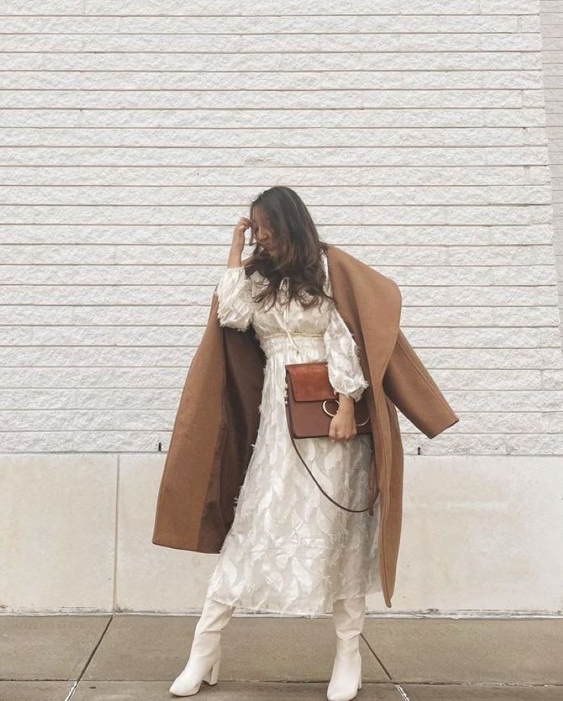 a woman in a burnout ivory dress with white boots and a camel coat and bag