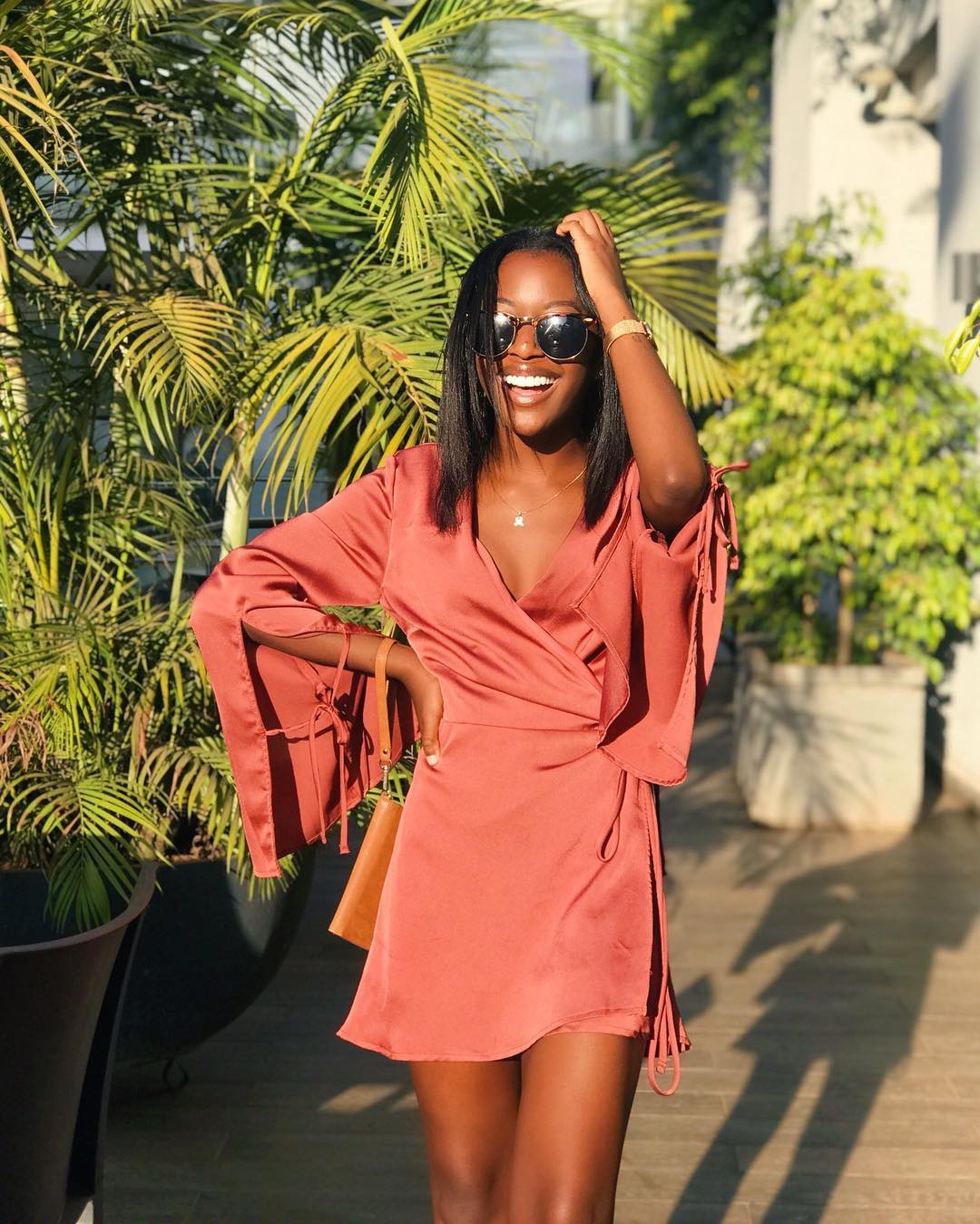 woman smiling in front of plants, wearing a coral dress and sunglasses