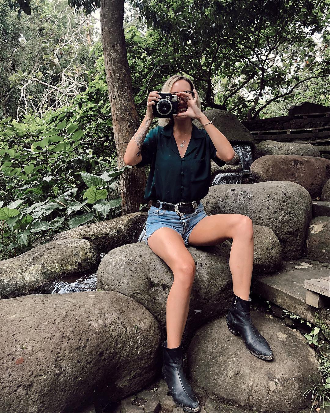 woman in denim shorts and button up taking pictures on a rock