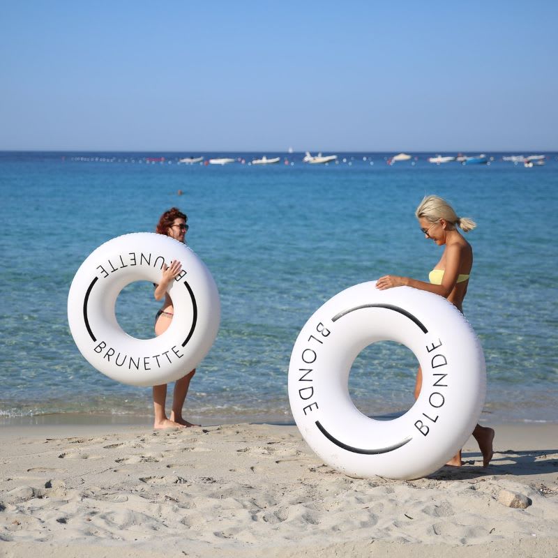 women walking on the beach with white round pool tubes saying blonde and brunette