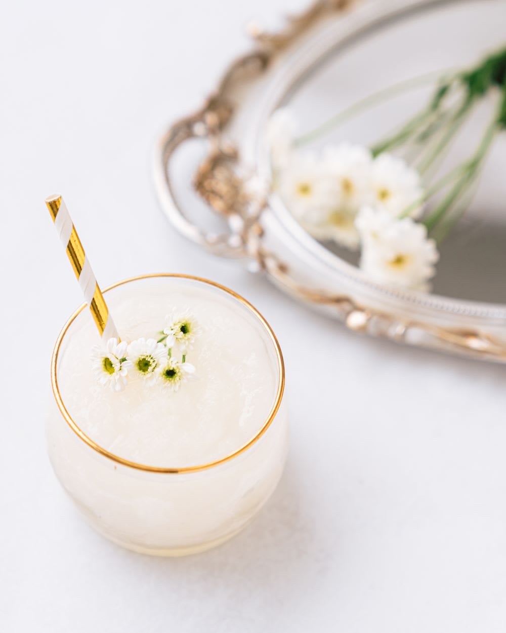 a glass with white wine slushie with daisies next to it on a tray and as garnish with gold striped straw