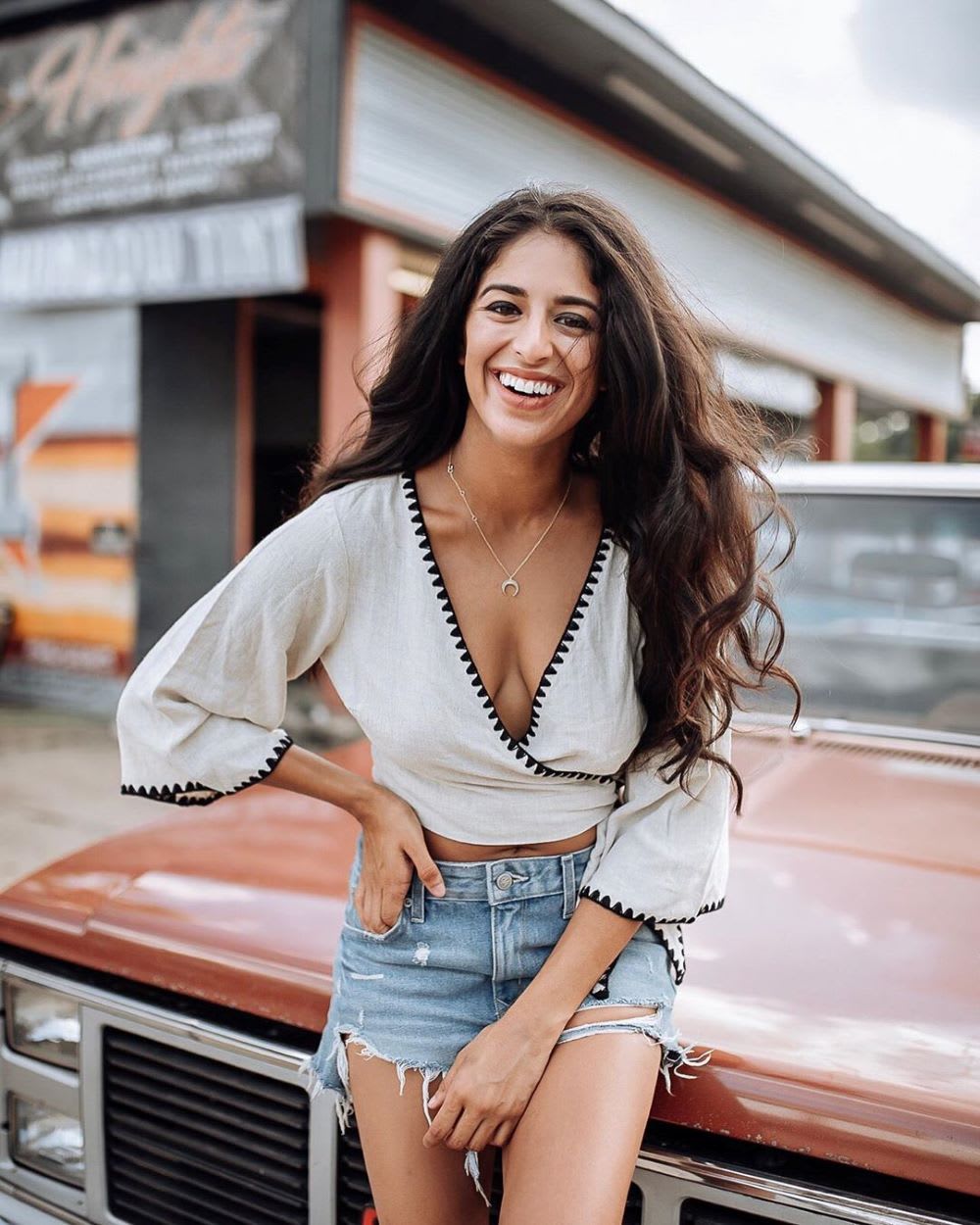 woman sitting smiling on the hood of a car in a crop top and denim shorts