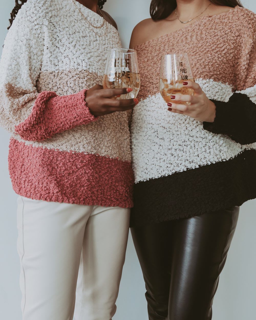 two woman from the neck down holding wine glasses and wearing similar color block sweaters off one shoulder