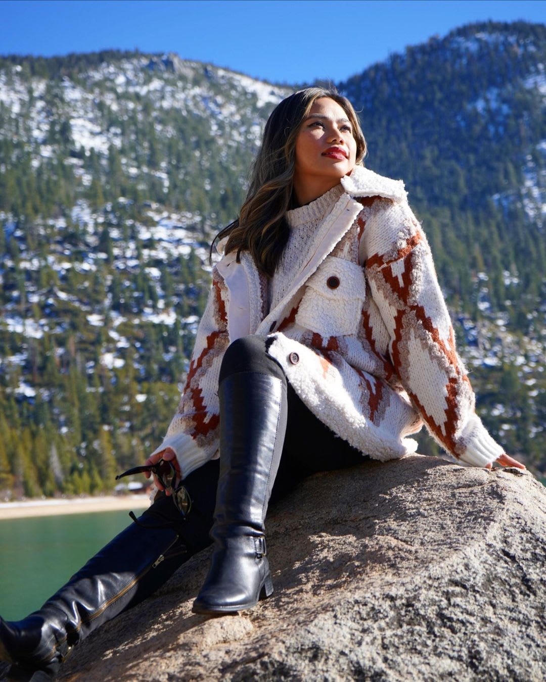 a woman wearing a printed cardigan sweater with black pants and tall black boots sitting on a rock by a mountainside lake