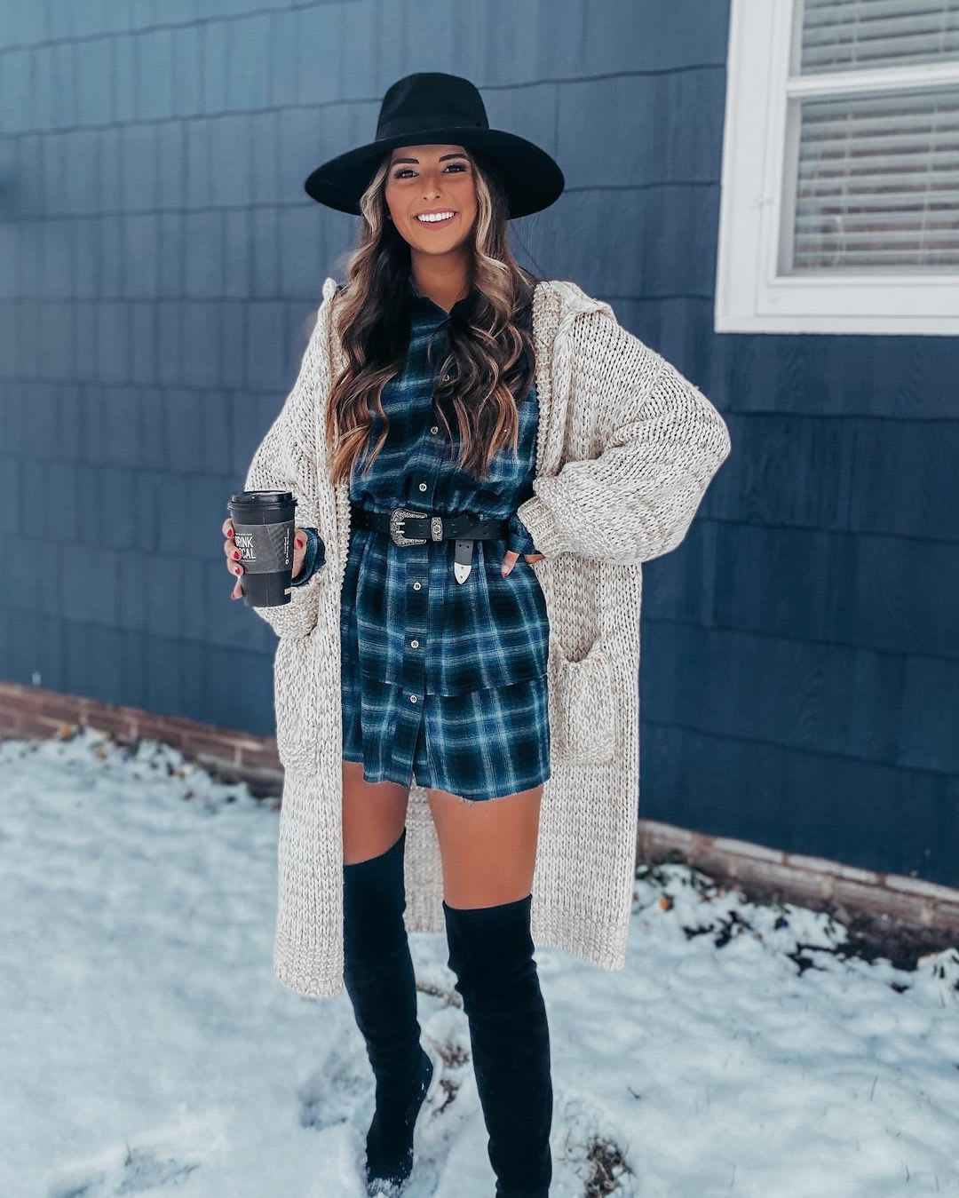a woman standing outside in the snow wearing a blue and black plaid shirtdress with black hat, belt, and otk boots with a cream cardigan over it