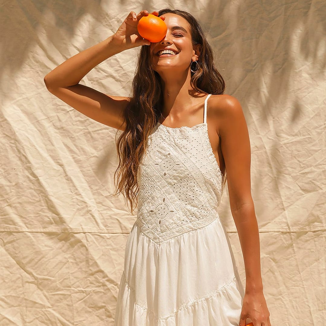 a model wearing a cream eyelet lace mini dress smiling toward the sun with an orange in her hand over her eye