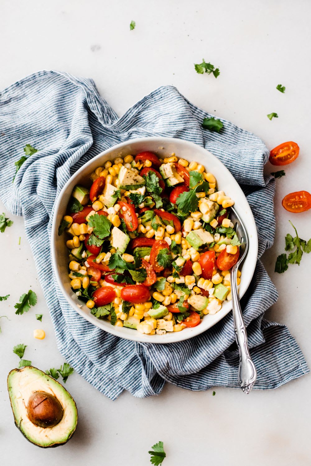 overhead shot of a bowl of corn tomato avocado salad with a napkin under it and a spoon and sliced avo next to it