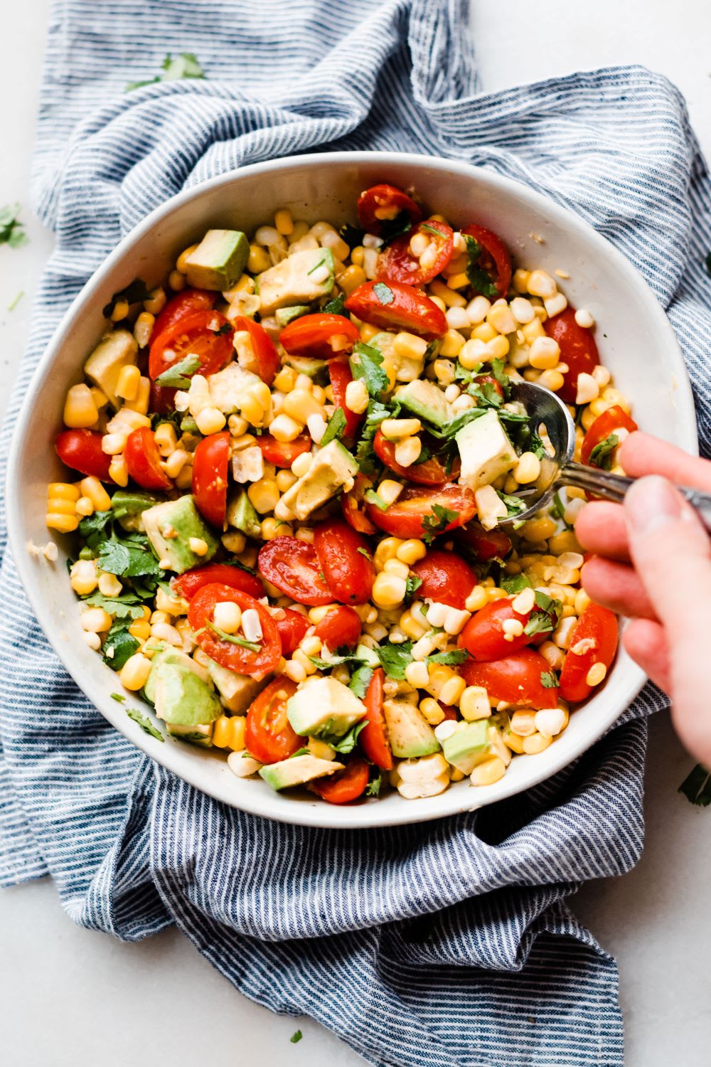 a hand with a spoon scooping from a bowl of corn tomato avocado salad