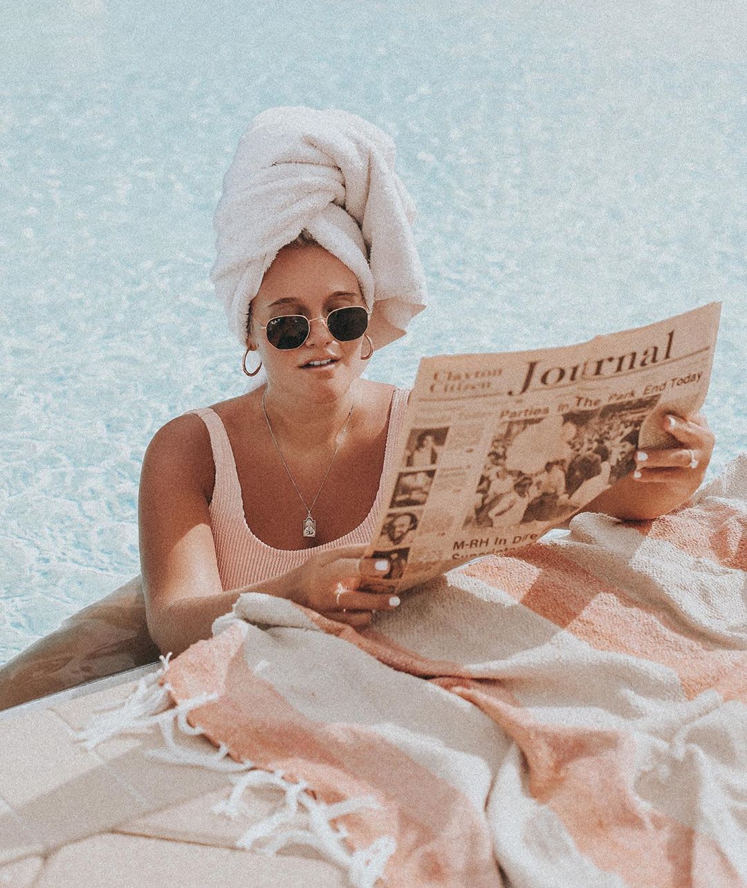 woman in a pool with a towel on her head reading a newspaper