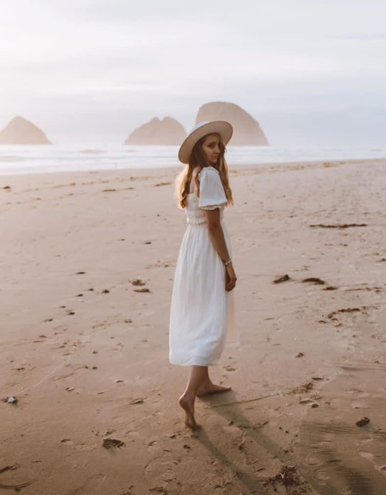 woman walking on a beach looking behind her wearing a white dress and hat