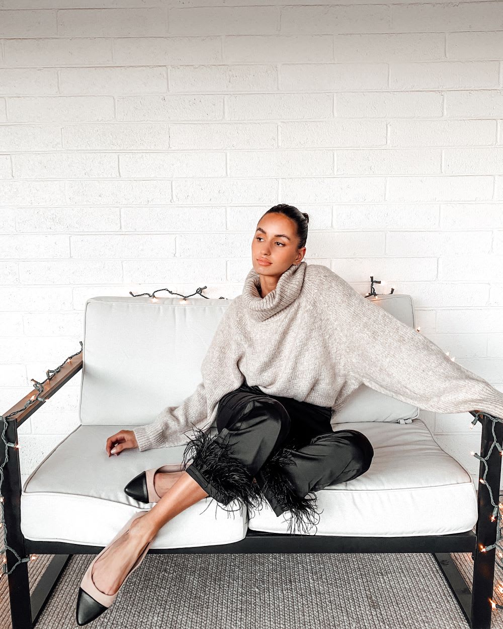 woman sitting on a couch wearing a beige oversized turtleneck sweater and black satin pants with feather trim and flats as a casual holiday outfits idea