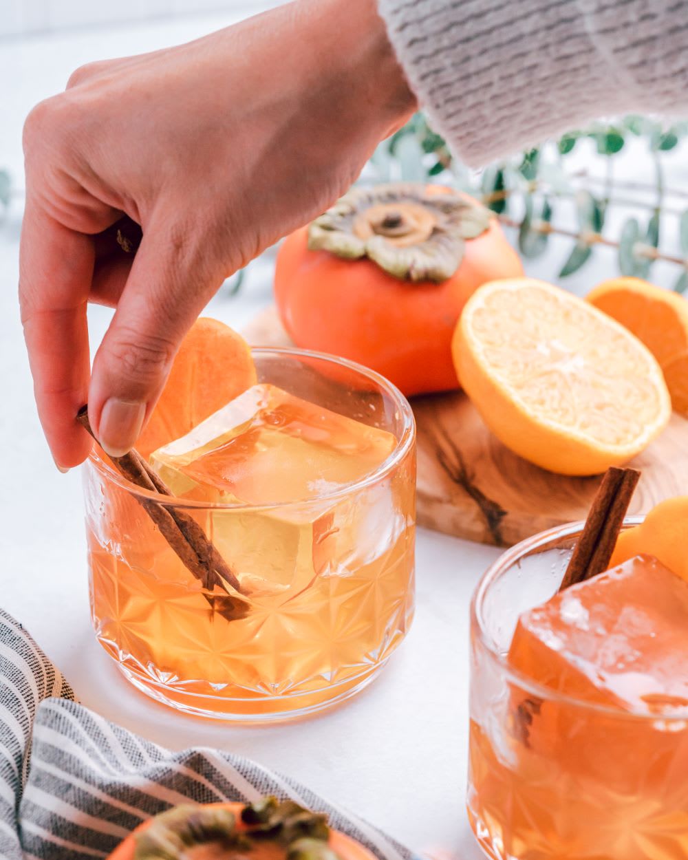 hand placing a cinnamon stick in an old fashioned cocktail recipe with persimmon and tangerine