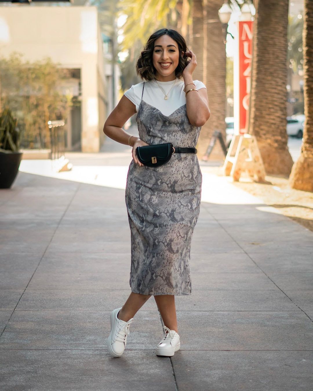 a woman wearing a snake print slip dress over a fitted white tee and a black belt bag shwing how to get a 90s look