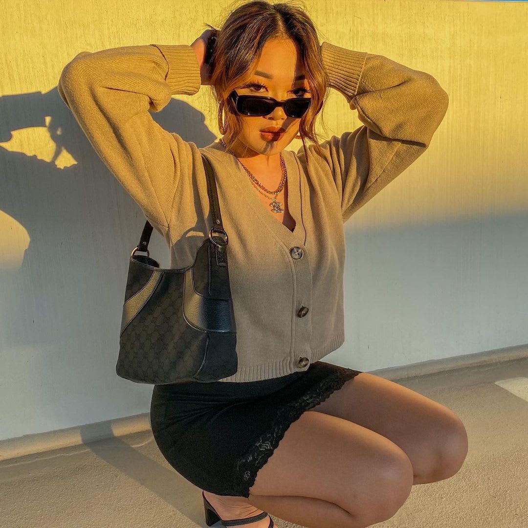 a woman crouching with her hands in her hair wearing a cropped cardigan and skinny sunglasses
