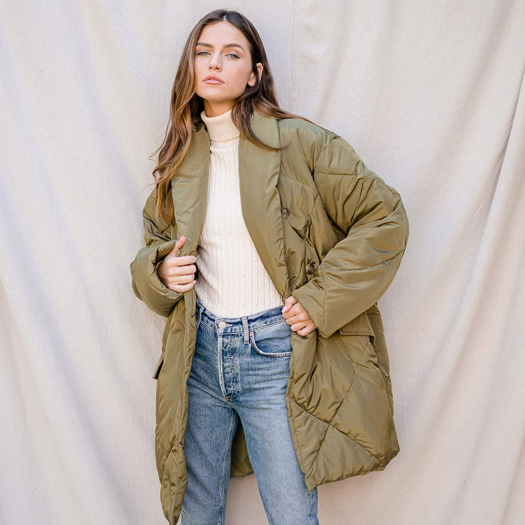 a woman standing in front of a curtain backdrop wearing an olive green quilted puffer coat with blue jeans and a white turtleneck