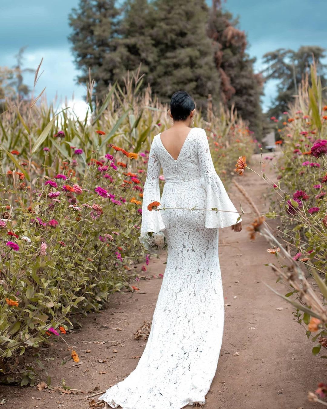 a bride wearing a lace lulus bridal gown walks down a flower path for a backyard wedding outdoors