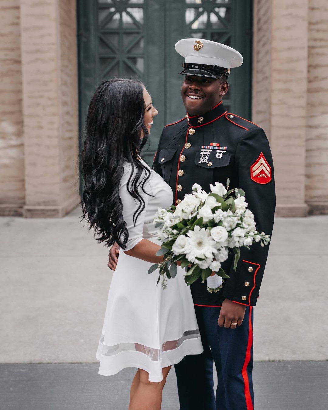 a bride in a little white dress and a groom in a military uniform smiling outside