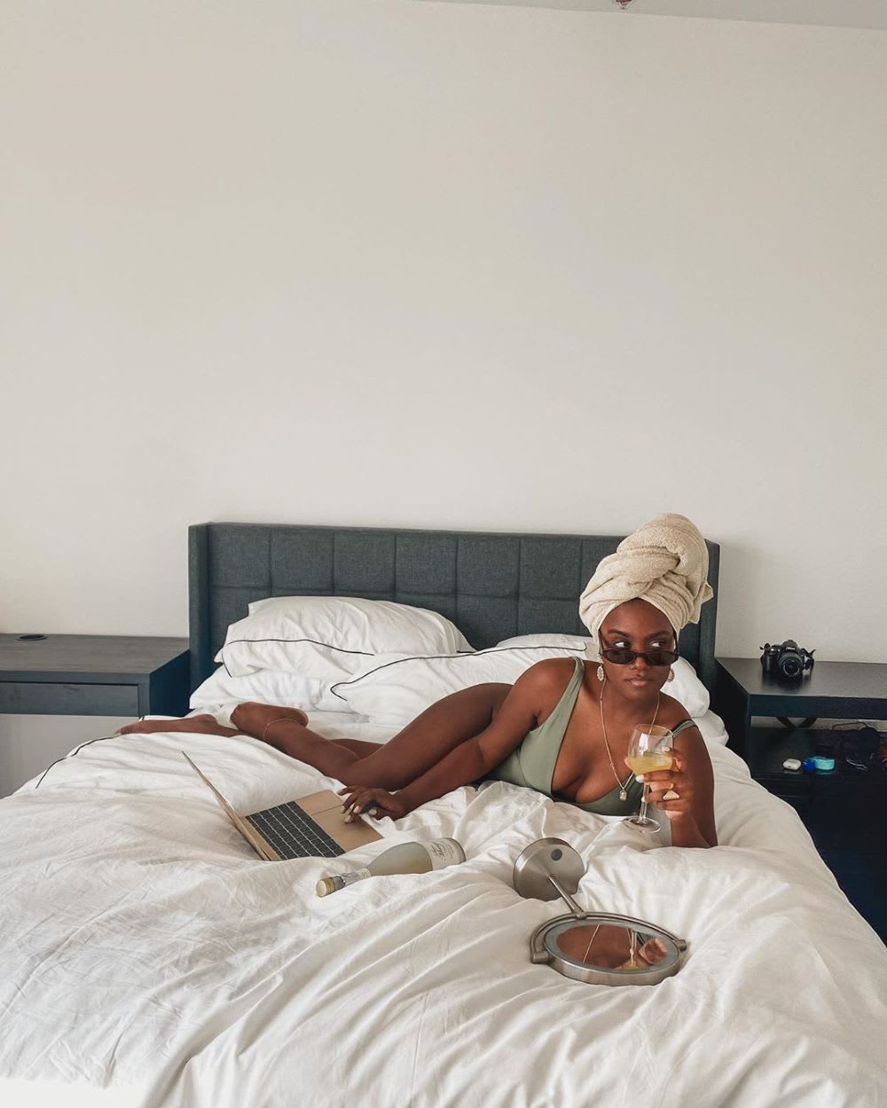 a woman on a bed wearing a bathing suit and towel on her head at her computer with a glass of wine