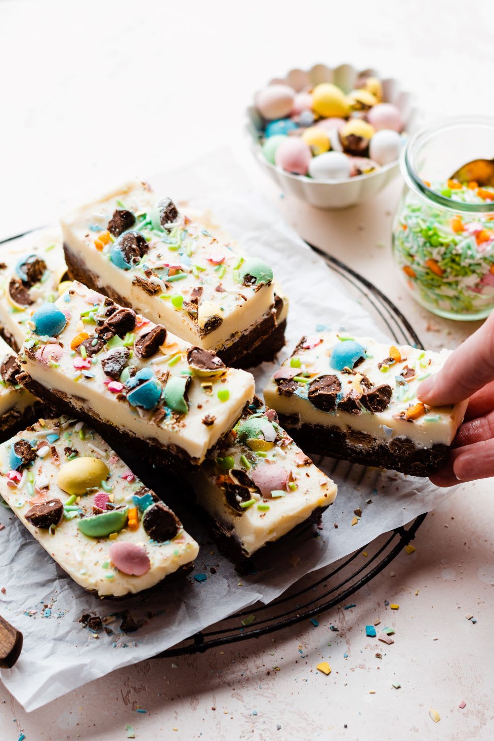 a hand taking a slice of double chocolate fudge with pastel easter candy eggs from a tray next to bowls of sprinkles and candy eggs