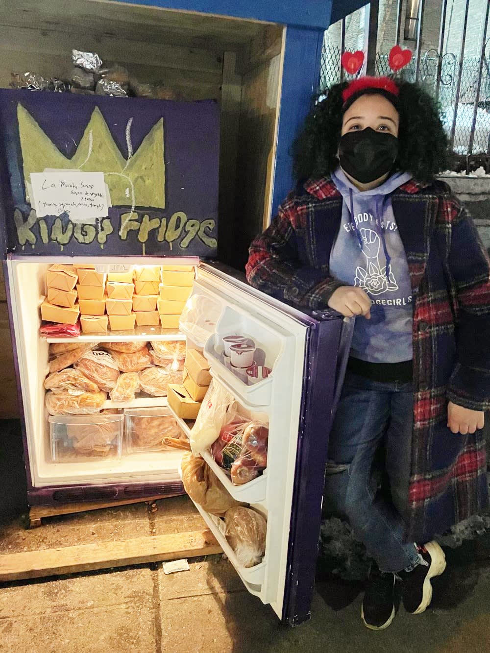 a woman poses with a community fridge full of food