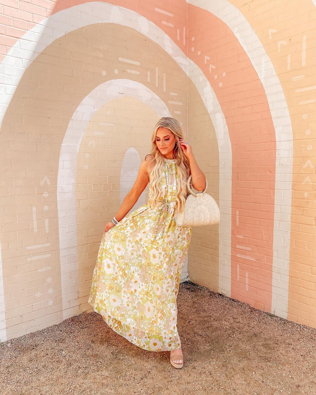 a woman wearing a 70s-inspired floral maxi dress with a woven straw bag in front of a tonal peach rainbow wall