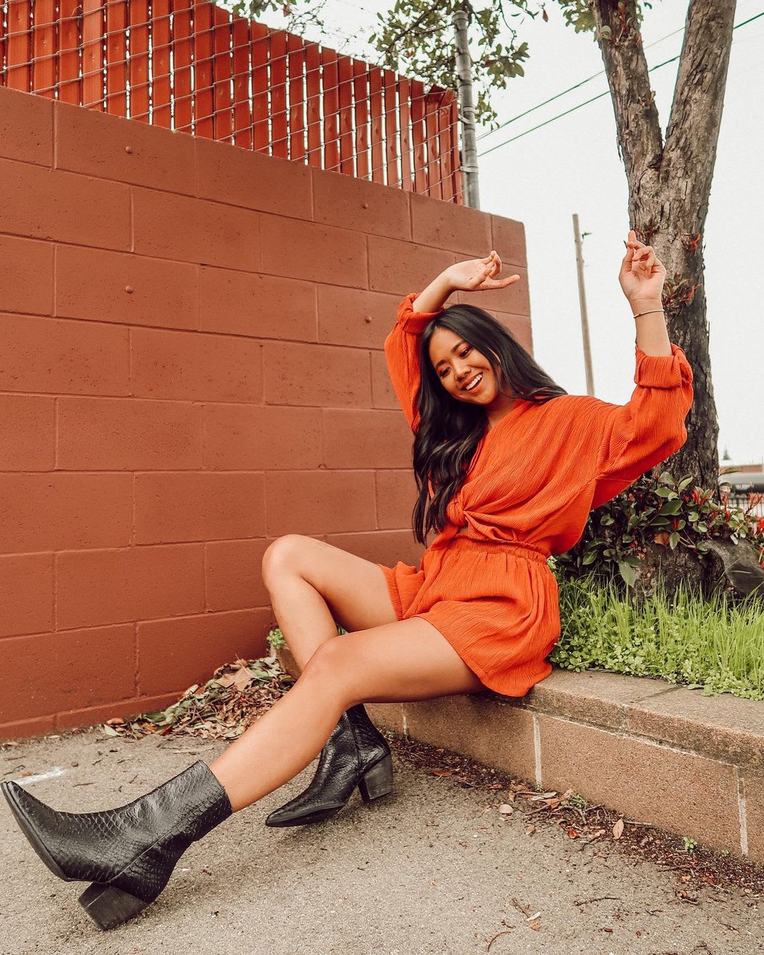 a woman in a rust red shorts and top with snake booties looking joyful sitting on a curb