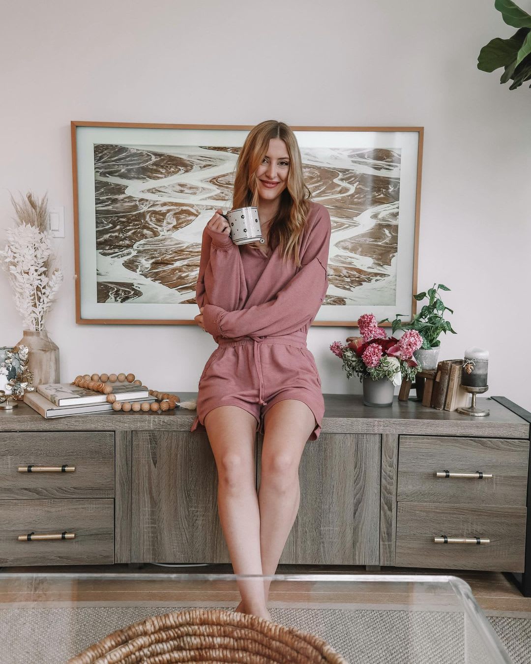 a woman in mauve loungewear set with a mug of tea sitting on her dresser smiling