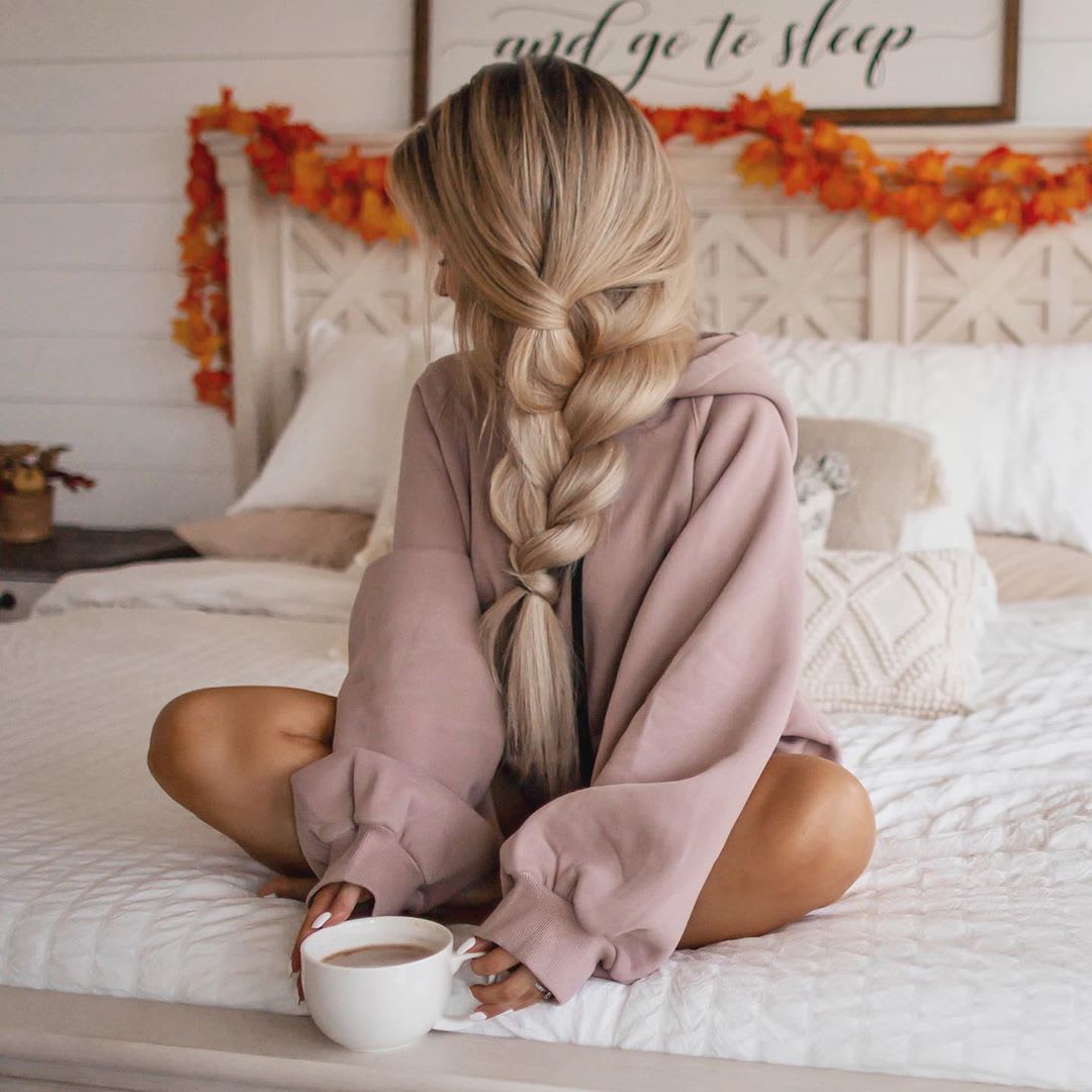 a woman sitting crosslegged on her made bed with a long side braid and holding a cup of coffee