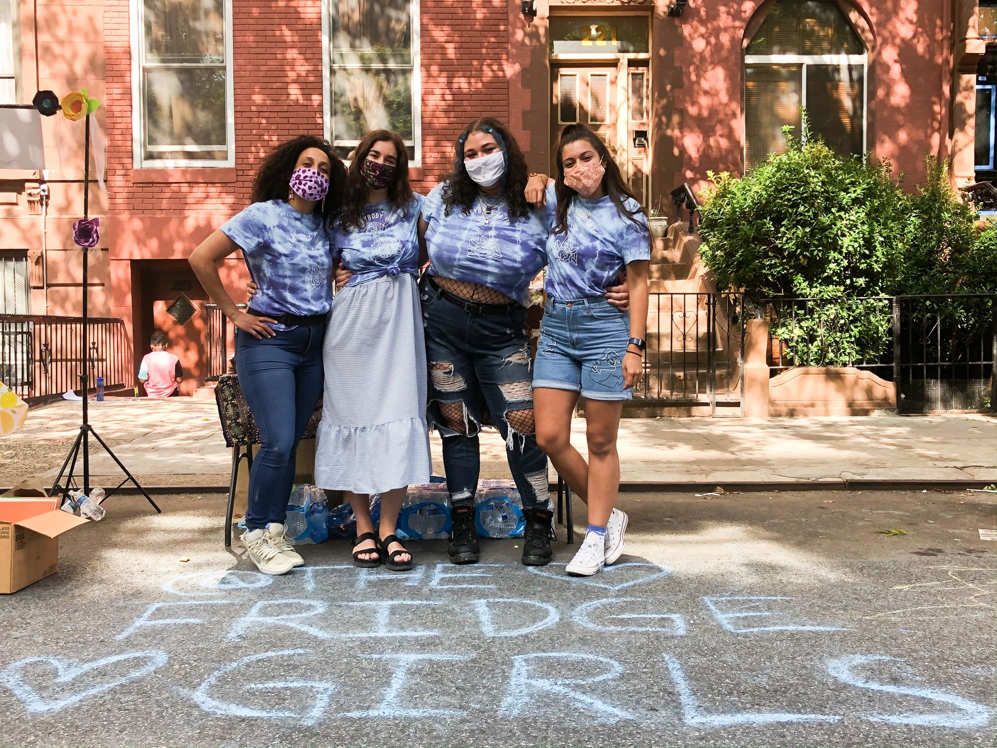 members of mutual aid community fridge organization The Fridge Girls pose in tie dye shirts and masks with their group name written in chalk on the street
