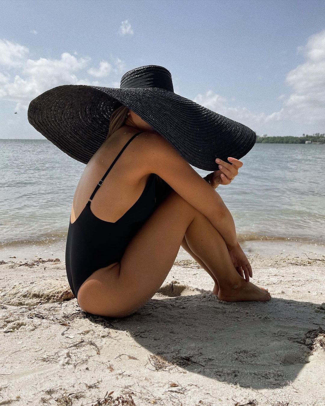 a woman sitting on a beach in a black one piece bathing suit with her arms across her knees and a black oversized black beach hat hiding her face
