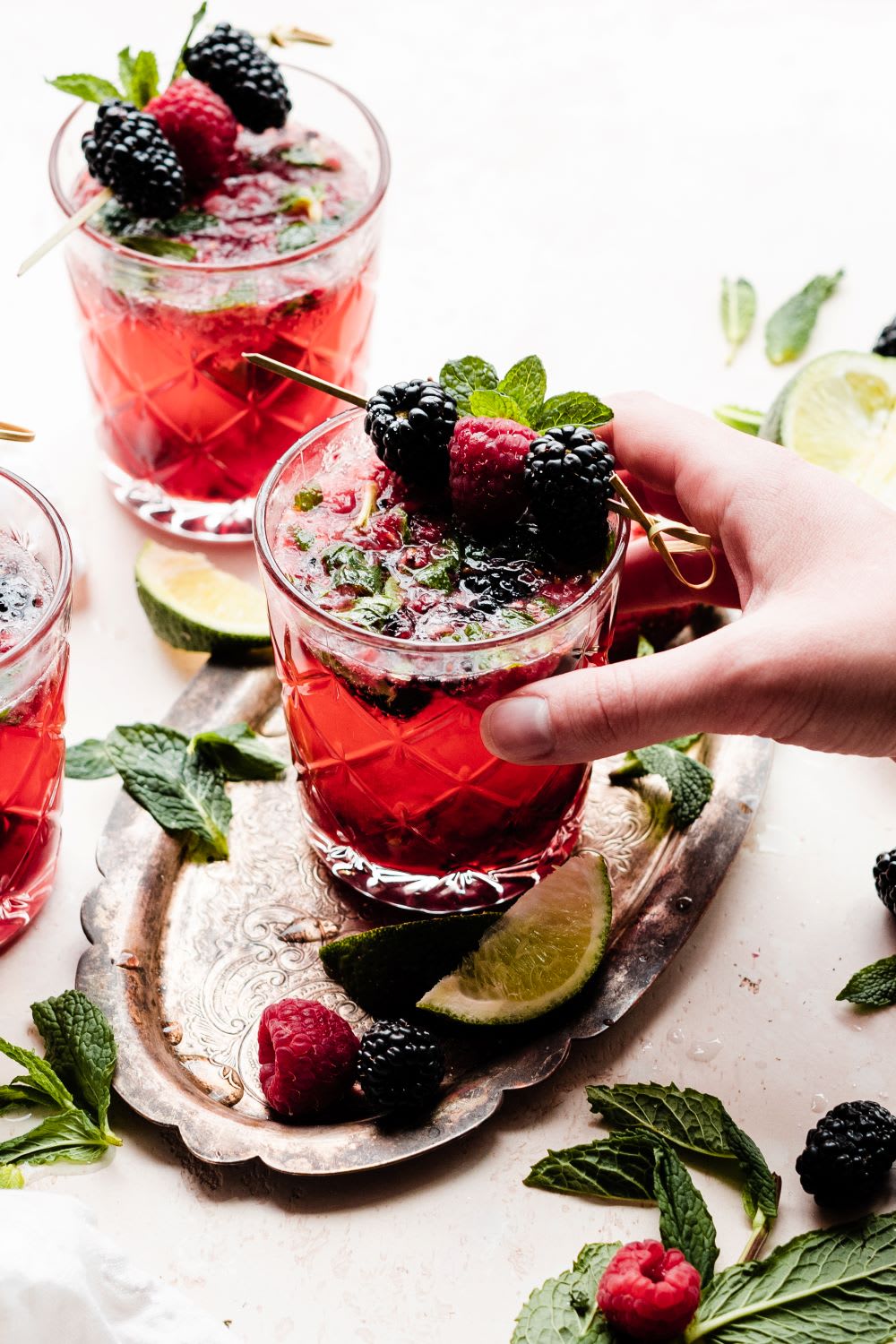 a hand picking up a glass of berry mojito with lime slices, raspberries, blackberries, and mint leaves scattered around 